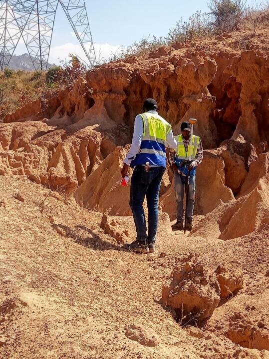 Two surveyors with a GPS pole in eroded ground below a transmission tower